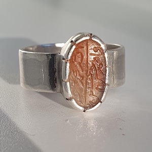 May include: A silver ring with an oval-shaped carnelian stone set in a silver bezel. The stone has an intricate design carved into it.
