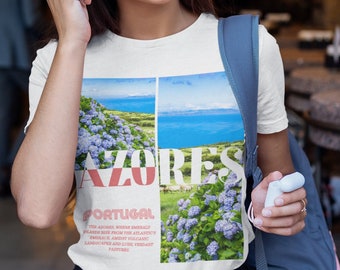 Op de afbeelding: Wit T-shirt met een grafische print van een landschap met blauwe bloemen en de tekst "AZORES PORTUGAL" De tekst onder de afbeelding luidt "De Azoren, waar smaragdgroene eilanden uit de omhelzing van de Atlantische Oceaan oprijzen, staan bekend om hun vulkanische landschappen en weelderige, groene weiden."