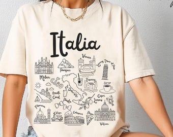 May include: Ivory-colored t-shirt with the word "Italia" in black script. The shirt features a detailed black and white illustration of Italian landmarks and phrases, including the Leaning Tower of Pisa and "Ciao".