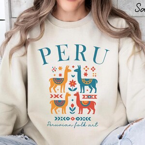 Peut inclure: Sweat-shirt couleur sable avec le mot "PERU" en lettres turquoise. En dessous, quatre illustrations colorées de lamas avec des motifs d'art populaire péruvien. Le texte "Peruvian folk art" est en dessous.