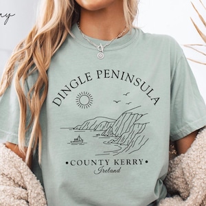 May include: A light green t-shirt with a black graphic of the Dingle Peninsula coastline, sun, and boat. The text "Dingle Peninsula County Kerry Ireland" is printed above and below the graphic. The shirt is made of a soft material.