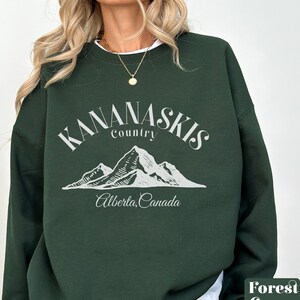 Pode incluir: Camisola verde floresta com uma estampa gráfica branca de montanhas e o texto "Kananaskis Country, Alberta, Canada".