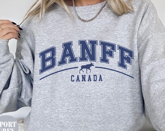 May include: A light gray sweatshirt with the word "BANFF" in navy blue arched lettering. Below the text is a moose graphic and the word "CANADA". The sweatshirt is labeled "SPORT GREY" in a small oval.