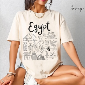May include: An ivory t-shirt featuring a black graphic print with the word Egypt at the top, surrounded by line art illustrations of Egyptian landmarks and symbols including the Great Pyramid of Giza, the Sphinx, a mummy, and the Eye of Horus.