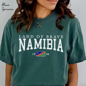 Peut inclure: T-shirt de couleur sarcelle avec l'inscription blanche "LAND OF BRAVE NAMIBIA". En dessous, un petit drapeau namibien et les chiffres "19 90". Les mots "Blue Spruce" sont dans le coin supérieur gauche.