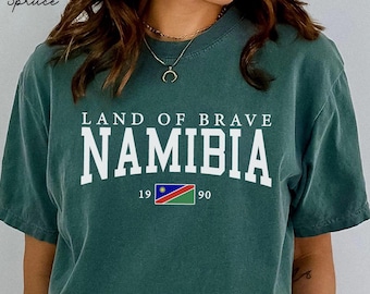 May include: A teal-colored t-shirt with the text "LAND OF BRAVE NAMIBIA" in white block letters. Below the text is a small Namibian flag and the numbers "19 90". The words "Blue Spruce" are in the upper left corner.
