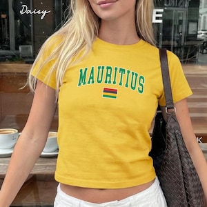 May include: A mustard yellow short-sleeved t-shirt featuring the word MAURITIUS in a green collegiate-style font arched over a small rectangular flag with horizontal stripes of red, blue, yellow, and green. The shirt is paired with white pants.