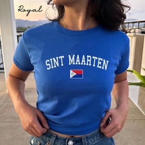 Può includere: T-shirt blu reale con la scritta bianca "SINT MAARTEN". Sotto il testo c'è una piccola bandiera di Sint Maarten. La maglietta a maniche corte è realizzata in materiale morbido. La parola "Royal" è scritta in corsivo.
