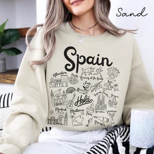 May include: Sand-colored sweatshirt with a black graphic of Spain landmarks and the word "Spain" in large script. The design includes illustrations of the Alhambra, paella, running of the bulls, and more. The word "Hola" is also included.
