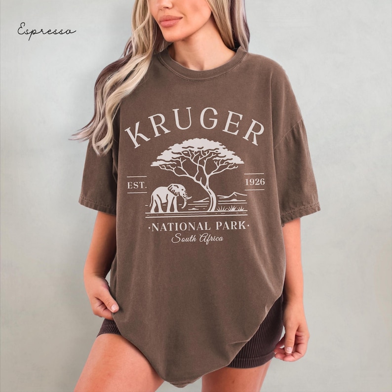 Puede incluir: Una camiseta marr&oacute;n con el texto "KRUGER NATIONAL PARK South Africa" en blanco. El dise&ntilde;o incluye un elefante, un &aacute;rbol y el a&ntilde;o 1926. La camiseta tiene mangas cortas y cuello redondo.
