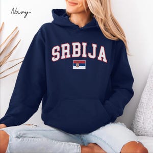 Puede incluir: Una sudadera con capucha azul marino que presenta la palabra SRBIJA en una fuente blanca arqueada con un contorno rojo. Debajo del texto hay una pequeña bandera serbia rectangular con franjas horizontales roja, azul y blanca y un escudo.