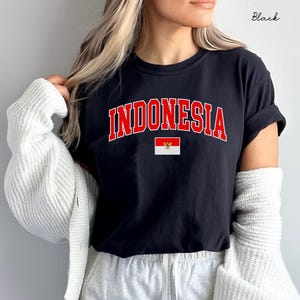May include: A black short-sleeved t-shirt featuring the word INDONESIA printed in a bold red arched font with a white outline. Below the text is a small rectangular graphic of the Indonesian flag with a central golden emblem.