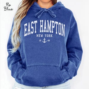 Pode incluir: Camisola azul com capuz com o texto branco "EAST HAMPTON NEW YORK". Abaixo do texto, há um desenho de âncora e os números "16 48". A camisola tem um bolso frontal e um capuz com cordão.