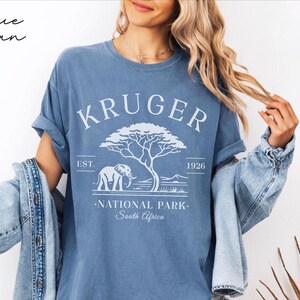 Puede incluir: Camiseta azul con el texto blanco "KRUGER NATIONAL PARK South Africa". El dise&ntilde;o incluye un elefante, un &aacute;rbol y el a&ntilde;o 1926. Las palabras "Blue Jean" est&aacute;n escritas en cursiva en la esquina superior izquierda.