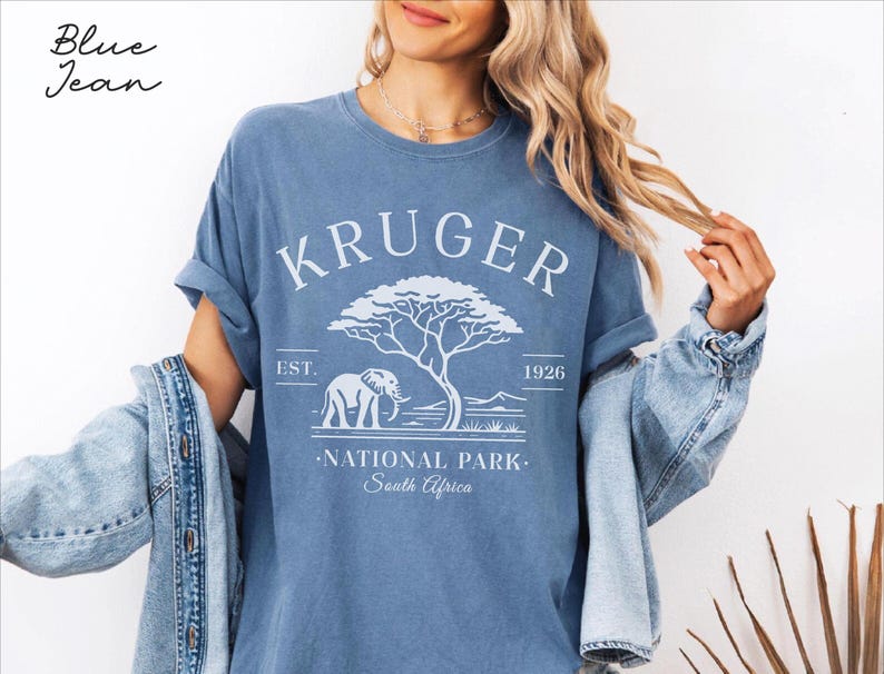 Puede incluir: Camiseta azul con el texto blanco "KRUGER NATIONAL PARK South Africa". El dise&ntilde;o incluye un elefante, un &aacute;rbol y el a&ntilde;o 1926. Las palabras "Blue Jean" est&aacute;n escritas en cursiva en la esquina superior izquierda.