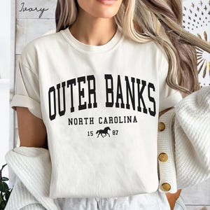 May include: Ivory-colored t-shirt with the text "Outer Banks, North Carolina" in bold black letters. Below the text is a silhouette of a horse and the numbers "15 87". The shirt has short sleeves.