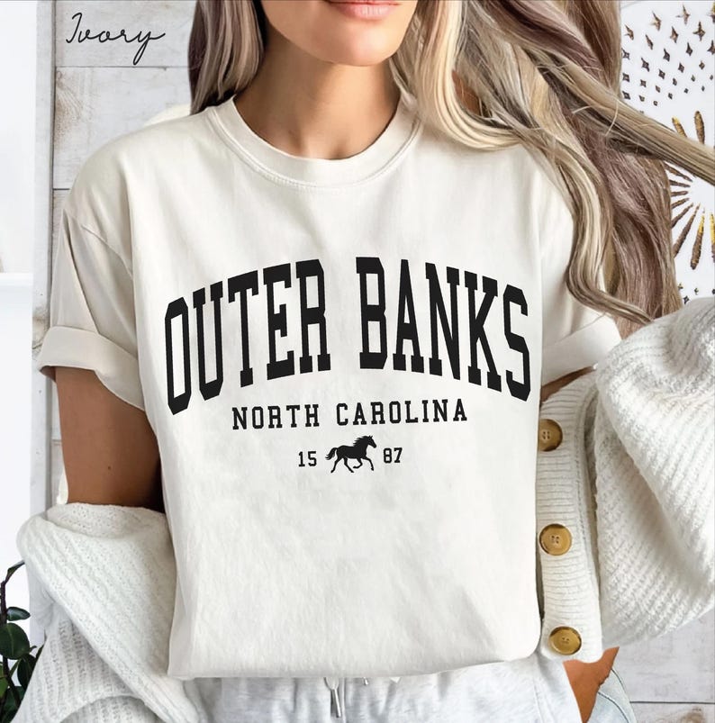 May include: Ivory-colored t-shirt with the text "Outer Banks, North Carolina" in bold black letters. Below the text is a silhouette of a horse and the numbers "15 87". The shirt has short sleeves.
