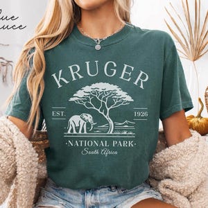 Puede incluir: Camiseta de color verde azulado con un dise&ntilde;o blanco. El dise&ntilde;o incluye el texto "KRUGER NATIONAL PARK South Africa", un elefante y un &aacute;rbol. El texto "EST. 1926" tambi&eacute;n forma parte del dise&ntilde;o.