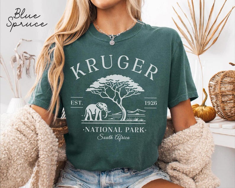 Puede incluir: Camiseta de color verde azulado con un dise&ntilde;o blanco. El dise&ntilde;o incluye el texto "KRUGER NATIONAL PARK South Africa", un elefante y un &aacute;rbol. El texto "EST. 1926" tambi&eacute;n forma parte del dise&ntilde;o.