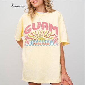 Könnte beinhalten: Ein hellgelbes Kurzarm-T-Shirt mit einem abgenutzten Grafikdruck. Das Design zeigt das Wort GUAM in großen rosa Buchstaben über einer stilisierten Sonne, Palmen und blauen Meereswellen mit dem darunter gedruckten Text Pacific Ocean.
