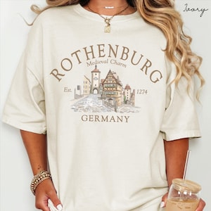 May include: An ivory short-sleeve t-shirt featuring a graphic print of a town scene with the text ROTHENBURG, Medieval Charm, Est. 1274, and GERMANY. The illustration depicts half-timbered buildings and a cobblestone street in brown and muted tones.