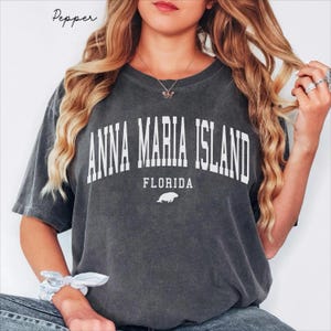 May include: Dark gray t-shirt with the text "ANNA MARIA ISLAND FLORIDA" in white block letters. A small white illustration of a manatee is below the text. The shirt has short sleeves and a crew neck.