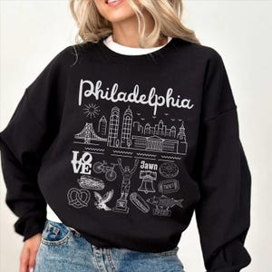 Könnte beinhalten: Ein schwarzes Sweatshirt mit Rundhalsausschnitt und weißem Grafikdruck. Das Design zeigt das Wort Philadelphia in Schreibschrift, eine Stadtsilhouette und verschiedene Symbole wie das LOVE-Zeichen, ein Fahrrad, einen Boxer, eine Glocke und einen Fisch.