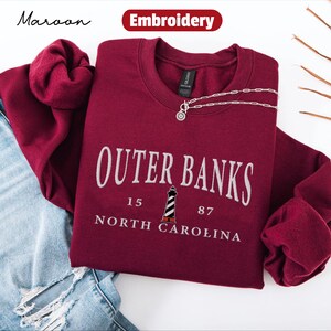 Pode incluir: Camisola bord&ocirc; com o texto "Outer Banks North Carolina" em branco, com um gr&aacute;fico de farol. Um colar de prata com um pingente repousa sobre a camisola. A palavra "Bordado" est&aacute; em uma faixa vermelha.