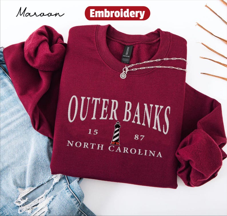 Pode incluir: Camisola bord&ocirc; com o texto "Outer Banks North Carolina" em branco, com um gr&aacute;fico de farol. Um colar de prata com um pingente repousa sobre a camisola. A palavra "Bordado" est&aacute; em uma faixa vermelha.