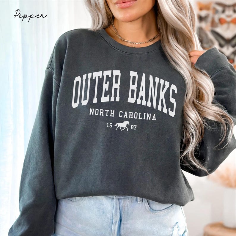 May include: Dark gray sweatshirt with the text "OUTER BANKS NORTH CAROLINA" in white block letters. Below the text is a small horse graphic and the numbers "15 87". The sweatshirt is a crew neck style.
