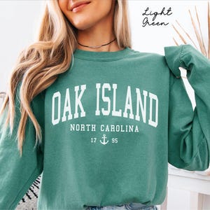 May include: A light green sweatshirt featuring the white text "OAK ISLAND NORTH CAROLINA". Below the text is "17 ⚓ 95". The sweatshirt is a solid colour and has long sleeves.