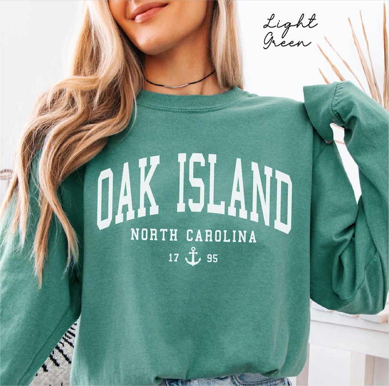 May include: A light green sweatshirt featuring the white text "OAK ISLAND NORTH CAROLINA". Below the text is "17 ⚓ 95". The sweatshirt is a solid colour and has long sleeves.