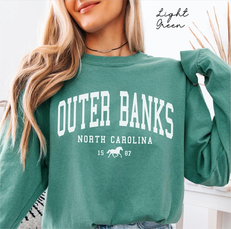May include: A light green sweatshirt with the text "OUTER BANKS NORTH CAROLINA" in white block letters. Below the text is a white horse graphic with the numbers "15 87". The words "Light Green" are written in cursive in the upper right corner.