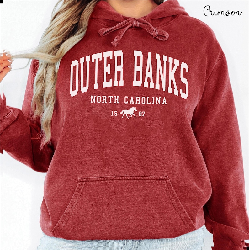May include: Crimson red Outer Banks hoodie with a drawstring hood and a kangaroo pocket. The white text reads "OUTER BANKS NORTH CAROLINA 15 87" with a horse graphic. A casual, comfortable sweatshirt.