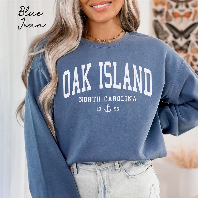 May include: A blue jean-coloured sweatshirt featuring the white text "OAK ISLAND NORTH CAROLINA". Below, the year "17 95" is displayed with an anchor symbol. The words "Blue Jean" are written in cursive in the upper left corner of the image.
