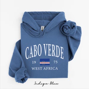 May include: An indigo blue hooded sweatshirt featuring the white text CABO VERDE in an arched font, the numbers 19 and 75, the national flag of Cape Verde, and the text WEST AFRICA printed in white on the center of the chest.