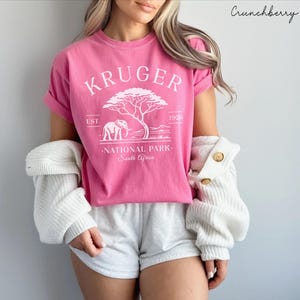 Puede incluir: Una camiseta rosa con un gr&aacute;fico blanco que dice "KRUGER NATIONAL PARK South Africa" y presenta una ilustraci&oacute;n de un &aacute;rbol y un elefante. La camiseta se usa con pantalones cortos grises y un su&eacute;ter blanco. El texto "Crunchberry" est&aacute; en la esquina superior derecha.