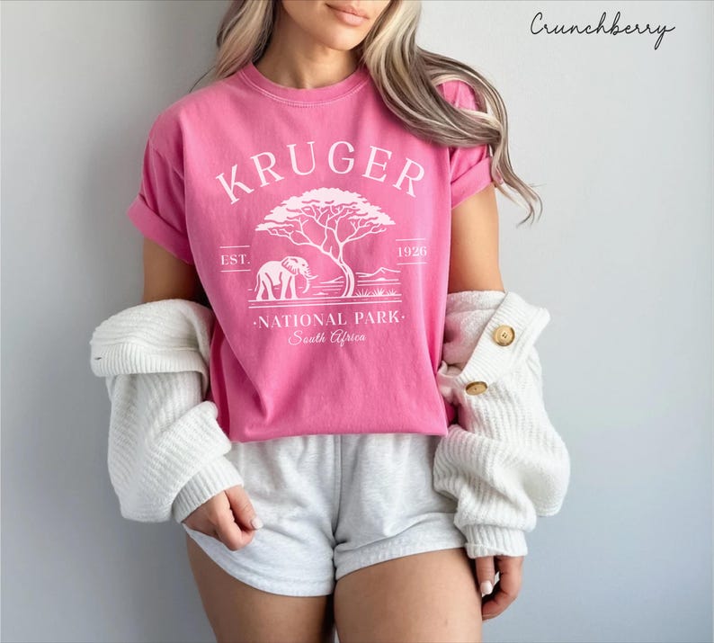 Puede incluir: Una camiseta rosa con un gr&aacute;fico blanco que dice "KRUGER NATIONAL PARK South Africa" y presenta una ilustraci&oacute;n de un &aacute;rbol y un elefante. La camiseta se usa con pantalones cortos grises y un su&eacute;ter blanco. El texto "Crunchberry" est&aacute; en la esquina superior derecha.