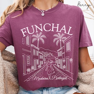 May include: Berry-colored t-shirt with a white graphic of Funchal, Madeira, Portugal. The design features a street scene with palm trees, buildings, and a person walking away. The text "FUNCHAL" is at the top.