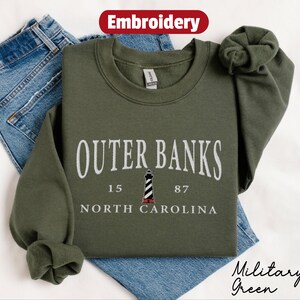 Pode incluir: Um moletom verde-oliva com o texto bordado "Outer Banks" acima de um gr&aacute;fico de farol e as palavras "North Carolina". O moletom est&aacute; dobrado em cima de um par de jeans azuis. A palavra "Bordado" est&aacute; em um oval vermelho.