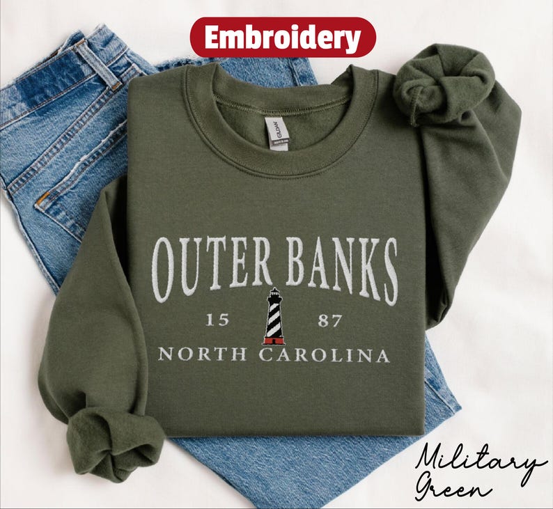 Pode incluir: Um moletom verde-oliva com o texto bordado "Outer Banks" acima de um gr&aacute;fico de farol e as palavras "North Carolina". O moletom est&aacute; dobrado em cima de um par de jeans azuis. A palavra "Bordado" est&aacute; em um oval vermelho.