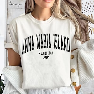 May include: Ivory-colored t-shirt with the text "ANNA MARIA ISLAND FLORIDA" in black, arched font. A small black illustration of a sea creature is below the word "FLORIDA". The shirt has short sleeves and a relaxed fit.