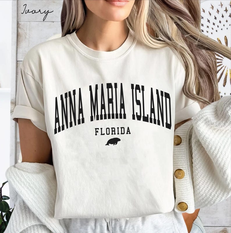May include: Ivory-colored t-shirt with the text "ANNA MARIA ISLAND FLORIDA" in black, arched font. A small black illustration of a sea creature is below the word "FLORIDA". The shirt has short sleeves and a relaxed fit.