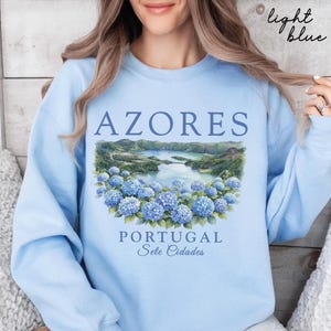 May include: A light blue crewneck sweatshirt featuring a graphic print of the Azores, Portugal, specifically Sete Cidades. The design includes blue hydrangea flowers in the foreground and a lake landscape, with the text Azores, Portugal, Sete Cidades.