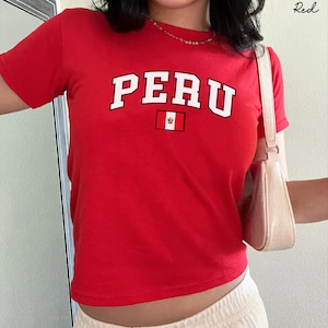 Op de afbeelding: Een felrood T-shirt met korte mouwen en het woord PERU in grote witte blokletters met een zwarte omlijning. Onder de tekst staat een kleine rechthoekige afbeelding van de Peruaanse vlag. Het T-shirt wordt gecombineerd met een lichtbeige schoudertas.