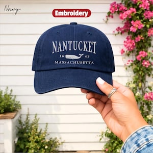 Könnte beinhalten: Marineblaue Baseballkappe mit dem Text "Nantucket Massachusetts" und einer Walgrafik. Die Kappe hat einen gebogenen Schirm und einen Knopf oben. Das Wort "Embroidery" steht in einem roten Banner.