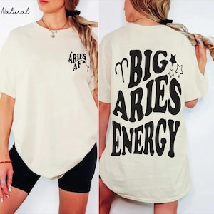 May include: A natural-colored t-shirt featuring black text. The front left chest displays the text ARIES AF with a small constellation graphic. The back features the large black text BIG ARIES ENERGY with an Aries symbol and star graphics.