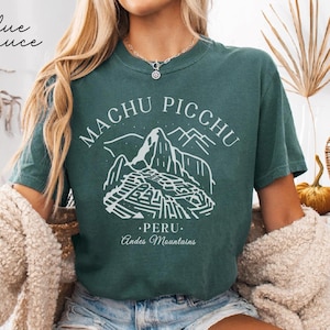 May include: Teal t-shirt with a white graphic of Machu Picchu and the text "Machu Picchu Peru Andes Mountains." The words "Blue Spruce" are written in cursive on the top left. The shirt is made of a soft material.