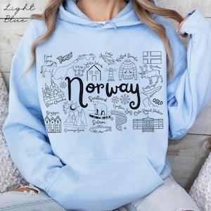 May include: Pale blue hoodie with a black graphic depicting the word "Norway" and Norwegian icons such as a reindeer, a polar bear, and a knitted sweater. The text "Light Blue" is at the top.