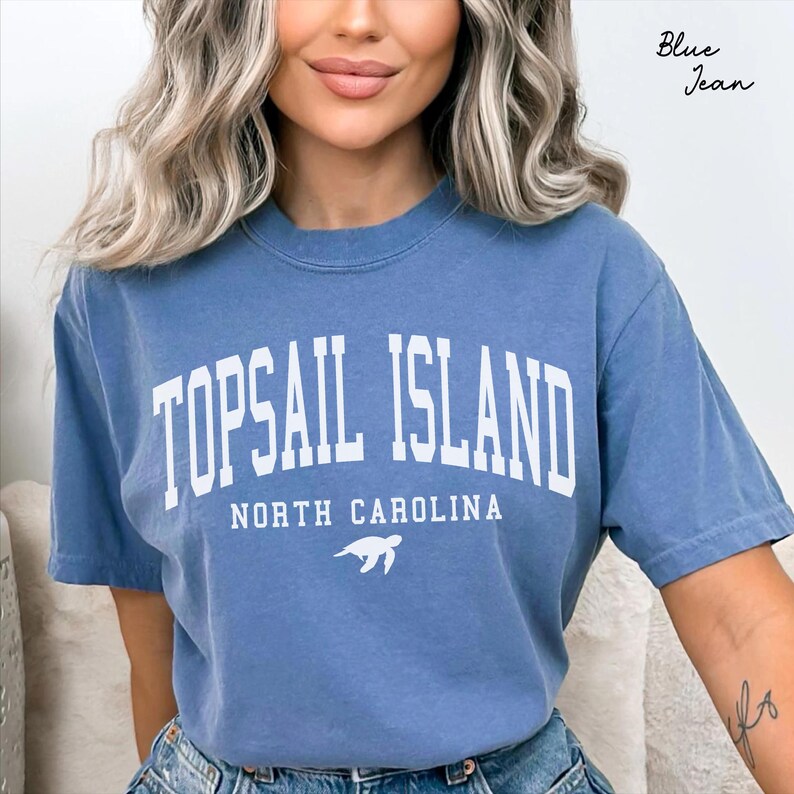 May include: A blue jean colored t-shirt with the text "TOPSAIL ISLAND NORTH CAROLINA" in white block letters. A white sea turtle graphic is below the text. The shirt has short sleeves and a crew neck.